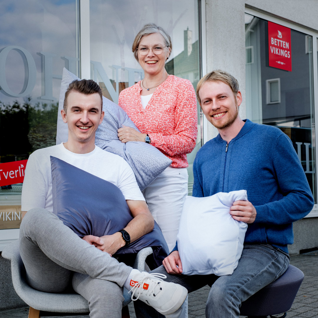 Zwei Mitarbeitende von Betten Vikings sitzen mit Kissen vor dem Geschäft, die Geschäftsführerin steht dahinter und lächelt